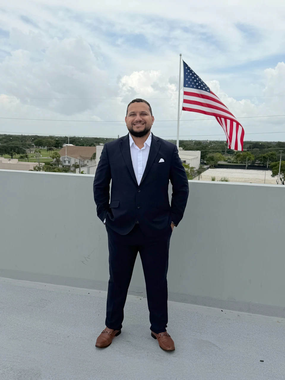 Roy Sánchez con traje azul y la bandera de Estados Unidos al fondo